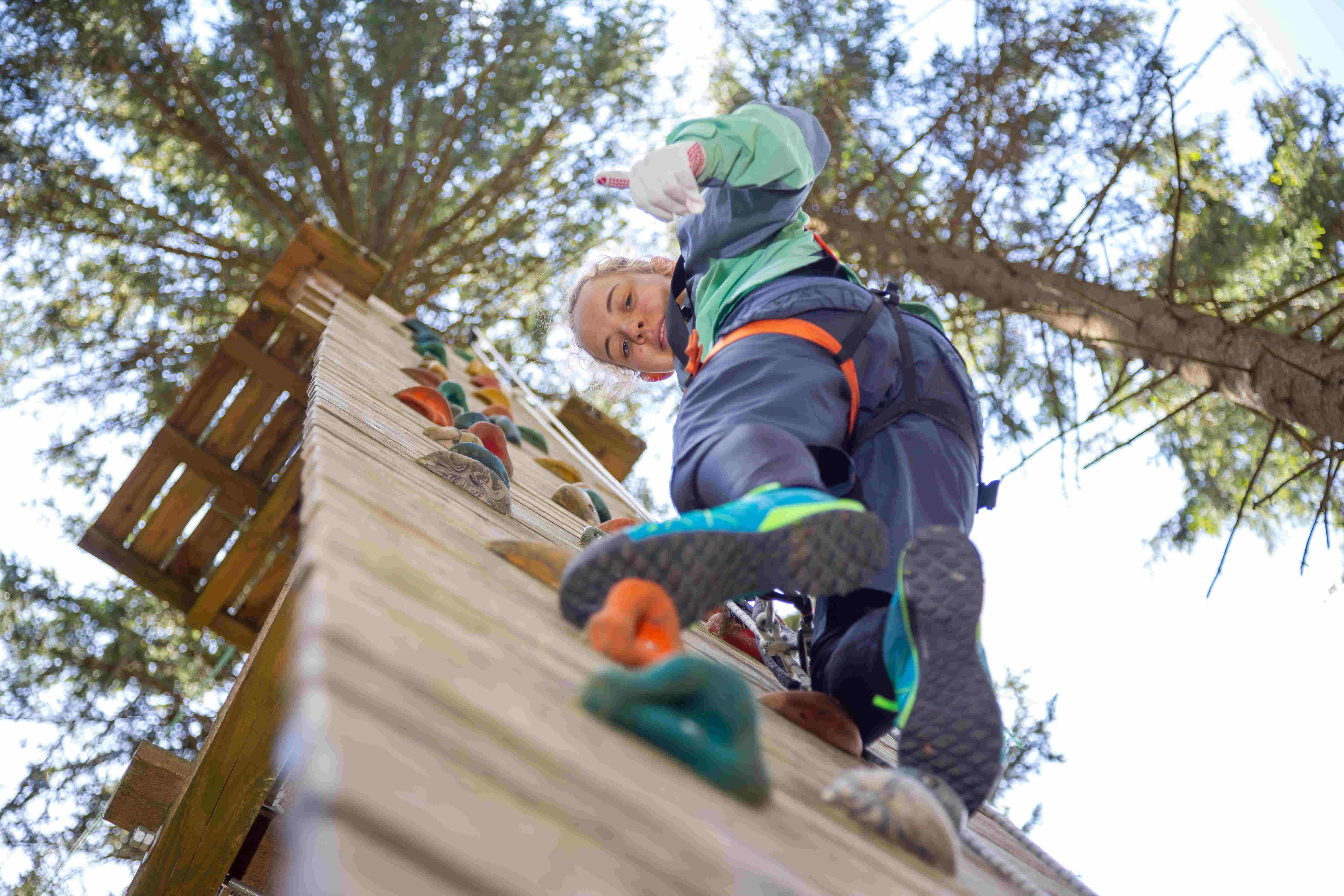 Sportpark Rabenberg: Kletterwald Abenteuerpark 860 Das Bild zeigt eine Frau, die eine Kletterwand erklimmt, aus einer niedrigen Perspektive. Die Frau ist im oberen Teil des Bildes zentriert und schaut direkt in die Kamera. Sie trägt eine grüne und graue Jacke, eine orangefarbene Sicherheitsausrüstung und blaue Schuhe mit gelben Akzenten. Ihre blonden Haare sind nach oben gebunden. Die Kletterwand ist aus Holz mit farbigen Klettergriffen. Der Hintergrund besteht aus Bäumen und blauem Himmel. Das Foto ist aus einem Winkel aufgenommen, der den Betrachter in die Lage der Person versetzt, die die Kletterwand hinaufschaut.