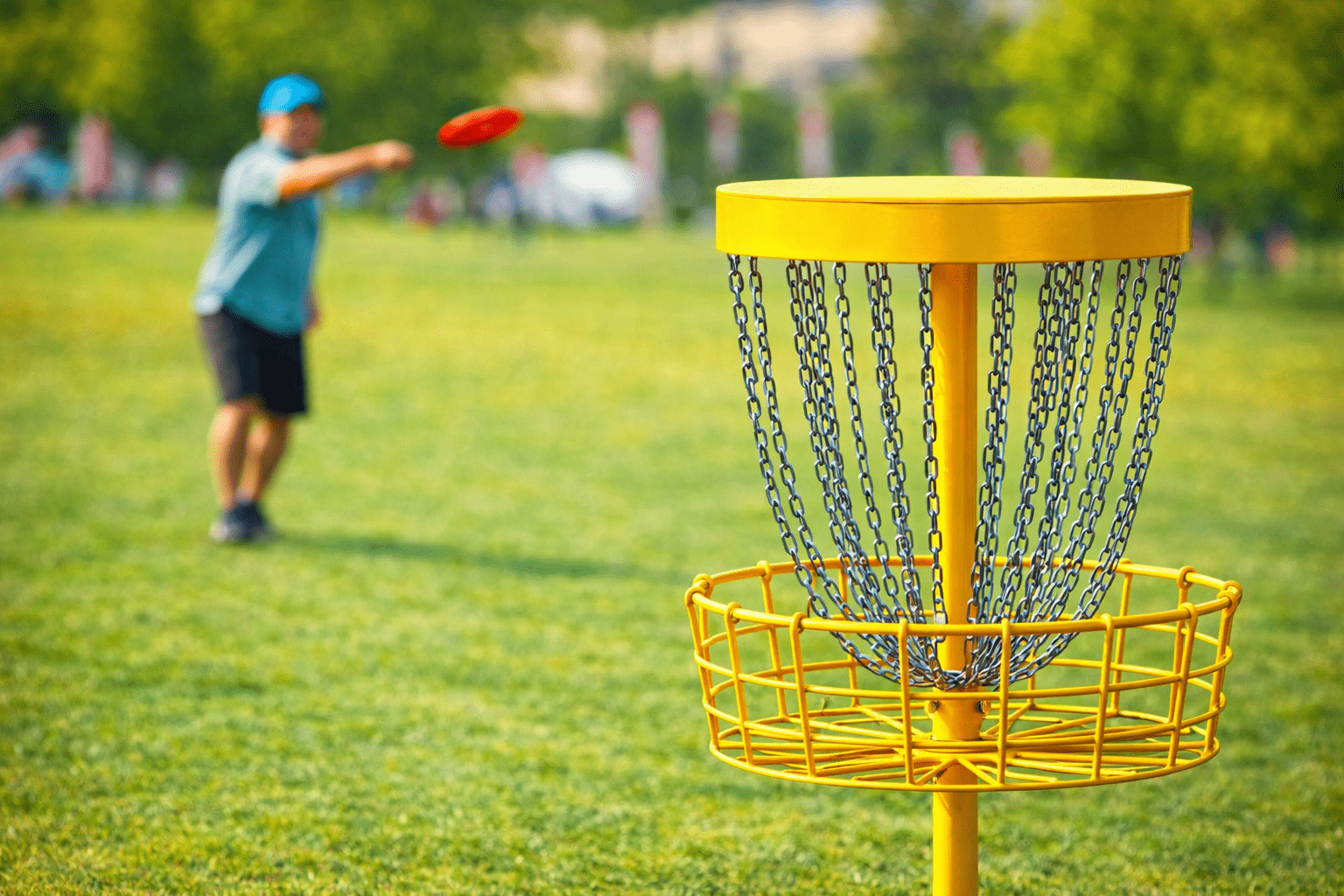 Rabenberg Sportkurse: Discgolf Parcours Das Bild zeigt eine Person, die eine Frisbeescheibe auf ein gelbes Frisbeegolf-Ziel wirft, das sich auf einem Rasen befindet. Die Person ist im Hintergrund unscharf, während das Ziel im Vordergrund scharf abgebildet ist. Die Frisbeescheibe ist rot und befindet sich in der Luft auf dem Weg zum Ziel.
