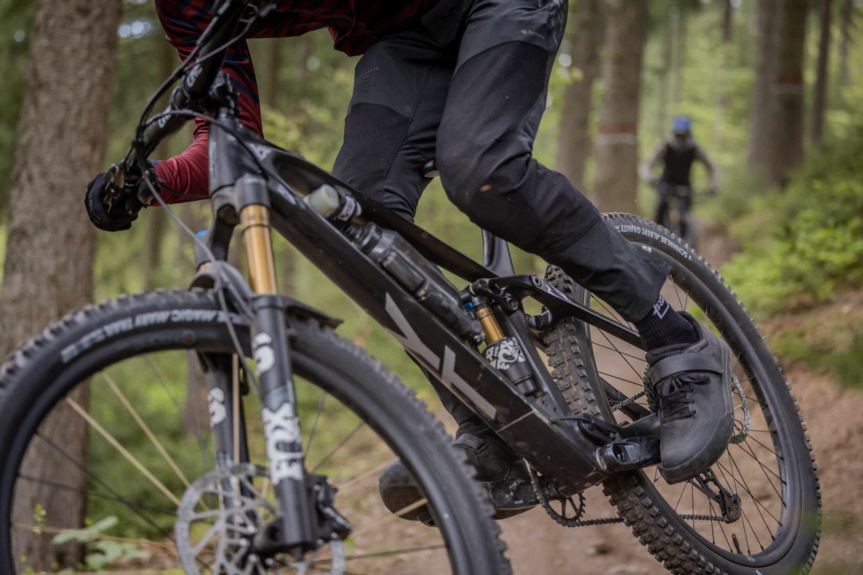 Rabenberg Sportkurse: Fahrtechnik Trail Das Bild zeigt einen Mountainbiker, der auf einem Waldweg fährt. Die Kamera befindet sich in niedriger Position und zeigt den Radfahrer und einen Teil des Fahrrads. Der Radfahrer trägt dunkle Kleidung und Handschuhe. Der vordere Teil des Fahrrads, einschließlich der Räder und der Federung, ist gut sichtbar. Im Hintergrund sind weitere Bäume und ein weiterer Radfahrer zu sehen, der in der Ferne fährt.