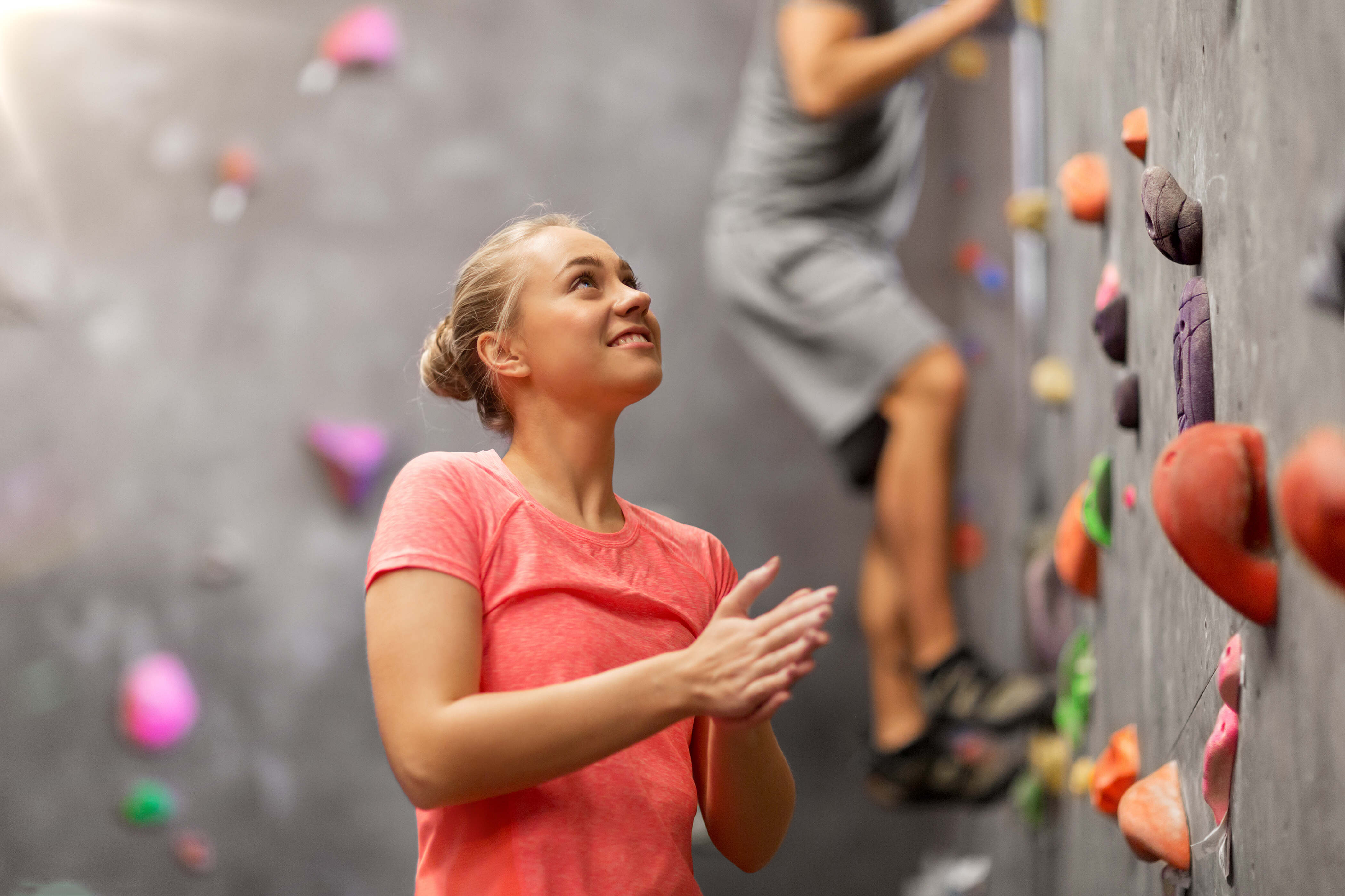Rabenberg Sportkurse: Indoorklettern Das Foto zeigt eine Frau vor einer Kletterwand. Sie ist blond und trägt ein rosa T-Shirt. Sie schaut nach oben und klatscht in die Hände. Im Hintergrund ist eine graue Kletterwand mit bunten Griffen zu sehen. Eine weitere Person klettert die Wand hinauf.
