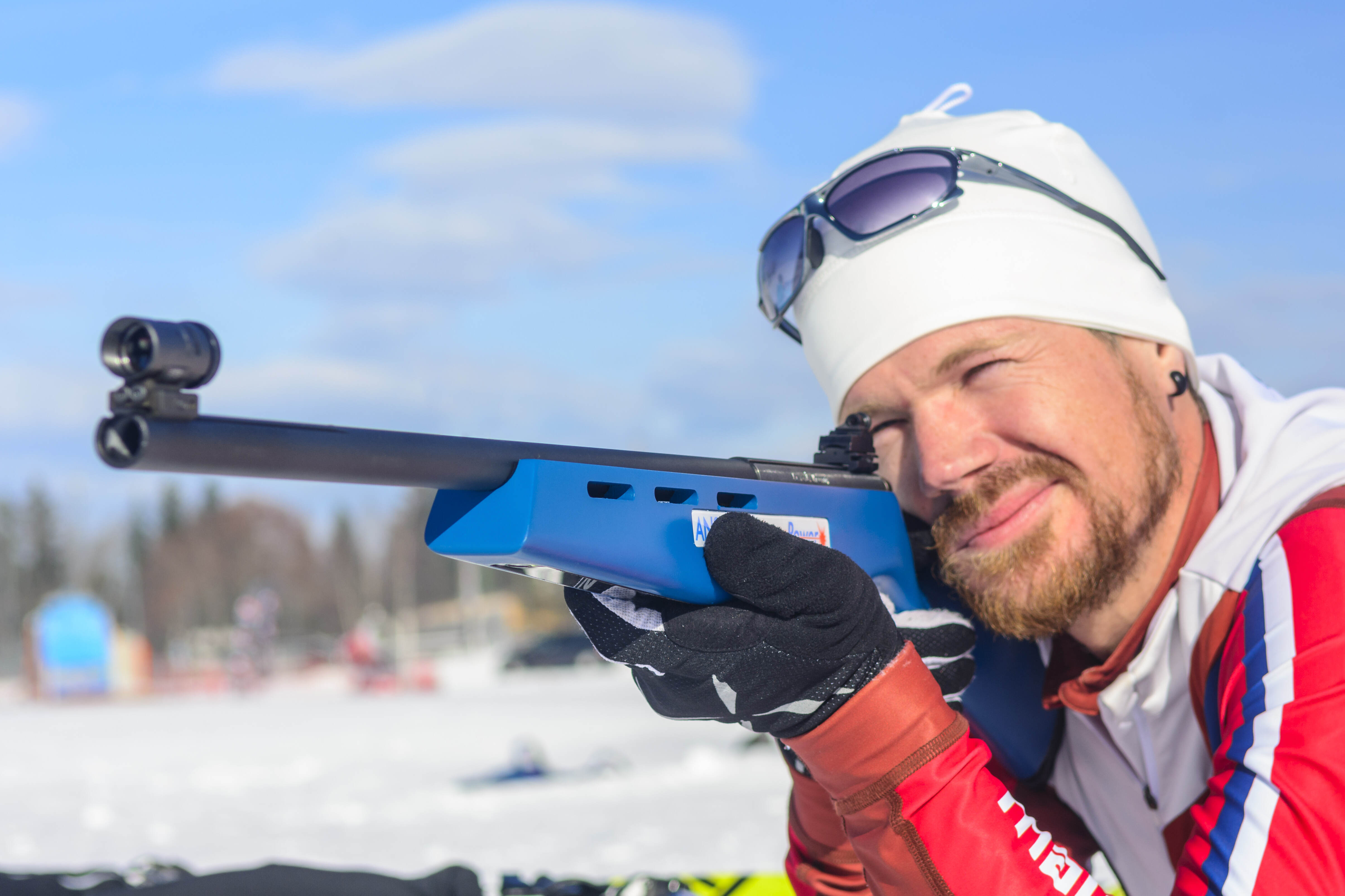 Rabenberg Sportkurse: Laserbiathlon Das Bild zeigt eine Person, die mit einem Gewehr zielt. Die Person trägt eine weiße Mütze, Sonnenbrille, eine rot-weiß-blaue Jacke und schwarze Handschuhe. Das Gewehr ist blau und schwarz. Im Hintergrund ist Schnee und ein blauer Himmel zu sehen.