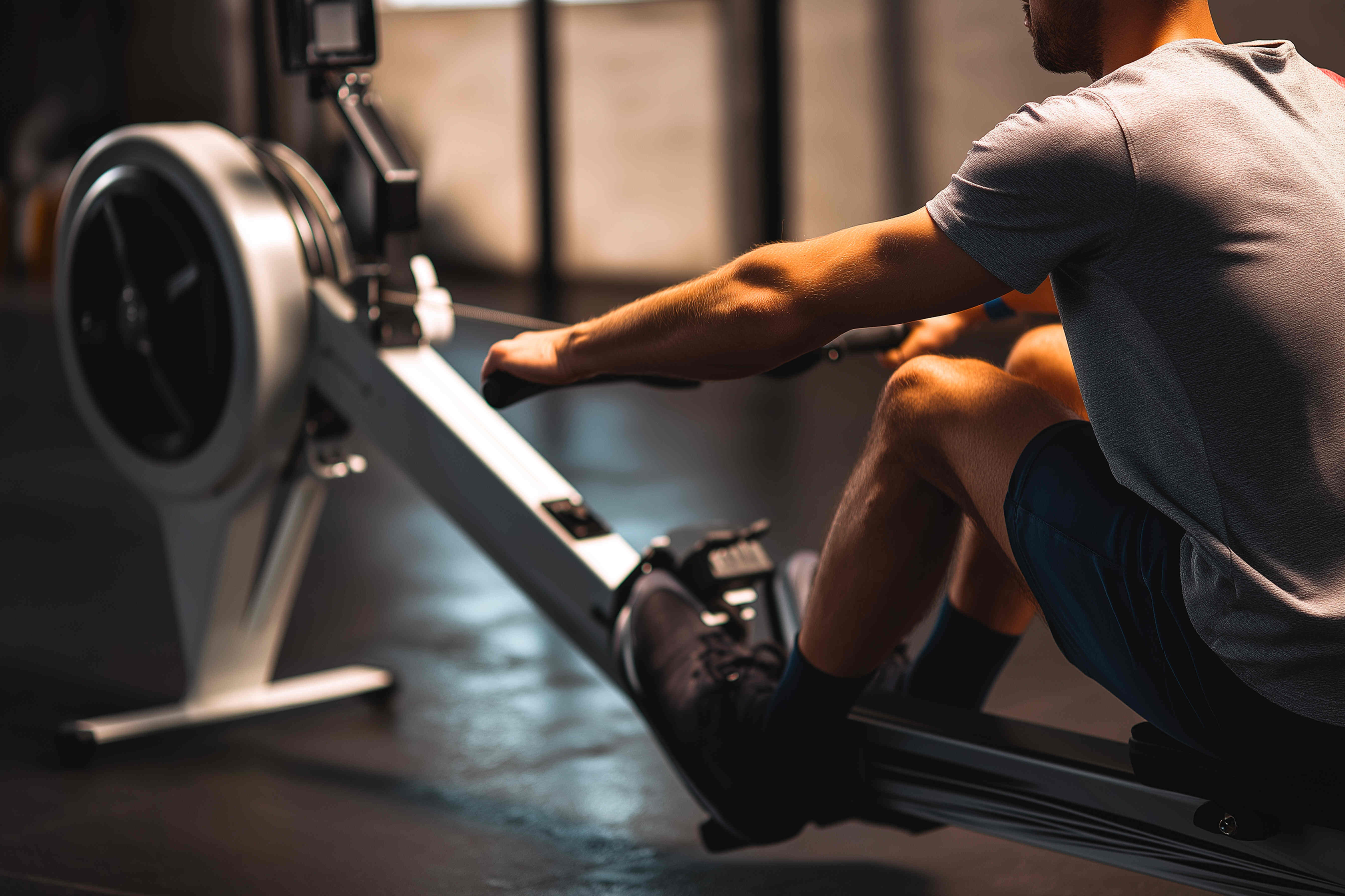 Rabenberg Sportkurse: Indoor Rowing Ein Mann trainiert auf einem Rudergerät. Nur sein Oberkörper und seine Beine sind im Bild zu sehen. Er trägt ein graues T-Shirt und blaue Shorts. Seine Hand hält den Griff des Rudergeräts. Seine Beine sind in den Fußstützen des Geräts fixiert. Das Rudergerät ist weiß und schwarz.