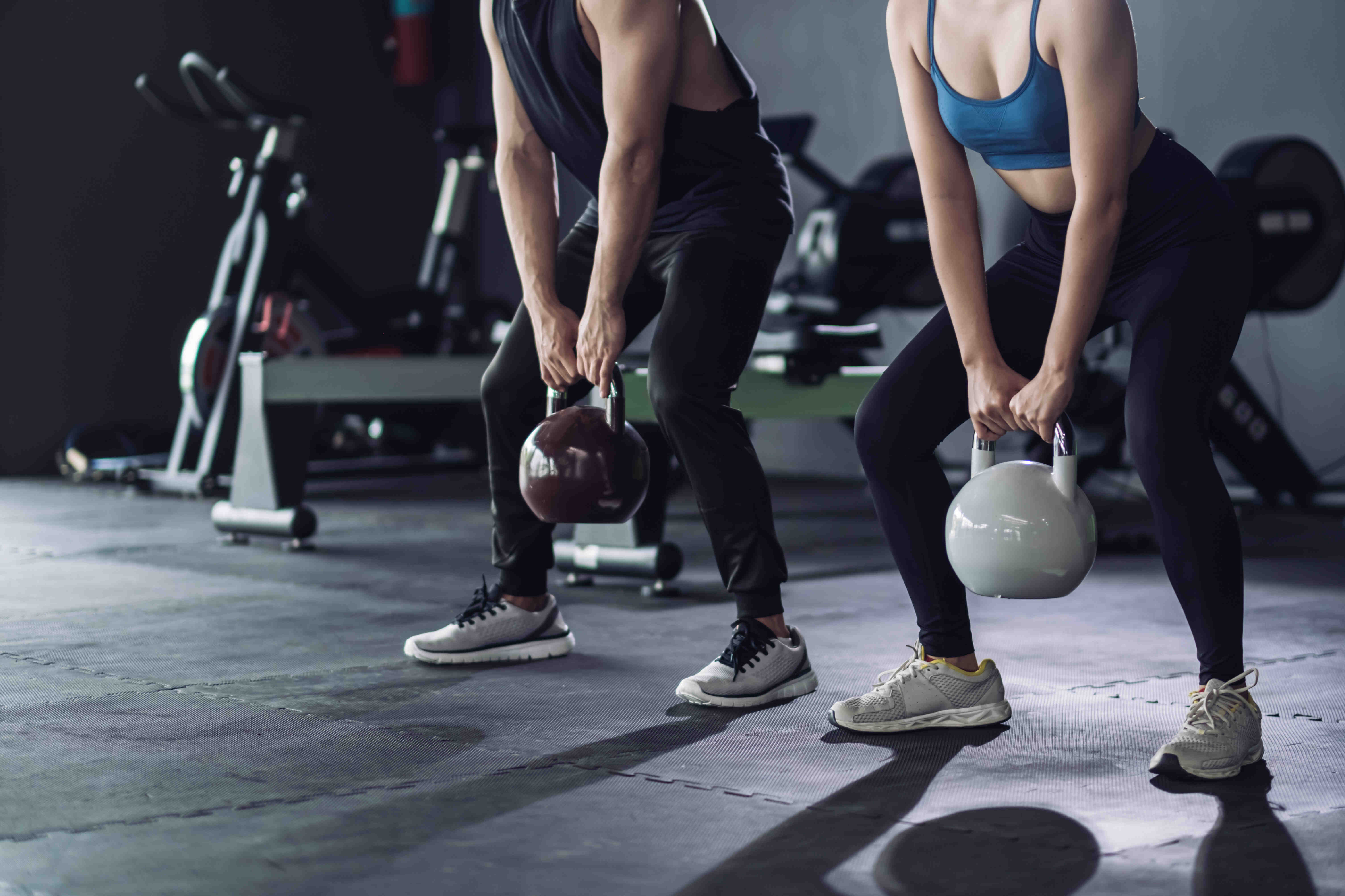 Rabenberg Sportkurse: Kettlebell Zwei Personen in Sportkleidung trainieren im Fitnessstudio. Sie stehen nebeneinander mit gebeugten Knien und halten jeweils eine Kettlebell. Die Person links ist männlich und trägt ein schwarzes Muskelshirt und eine schwarze Hose. Die Kettlebell ist dunkelbraun. Die Person rechts ist weiblich und trägt ein blaues Sport-BH und eine schwarze Hose. Ihre Kettlebell ist weiß. Im Hintergrund sind Fitnessgeräte zu sehen.
