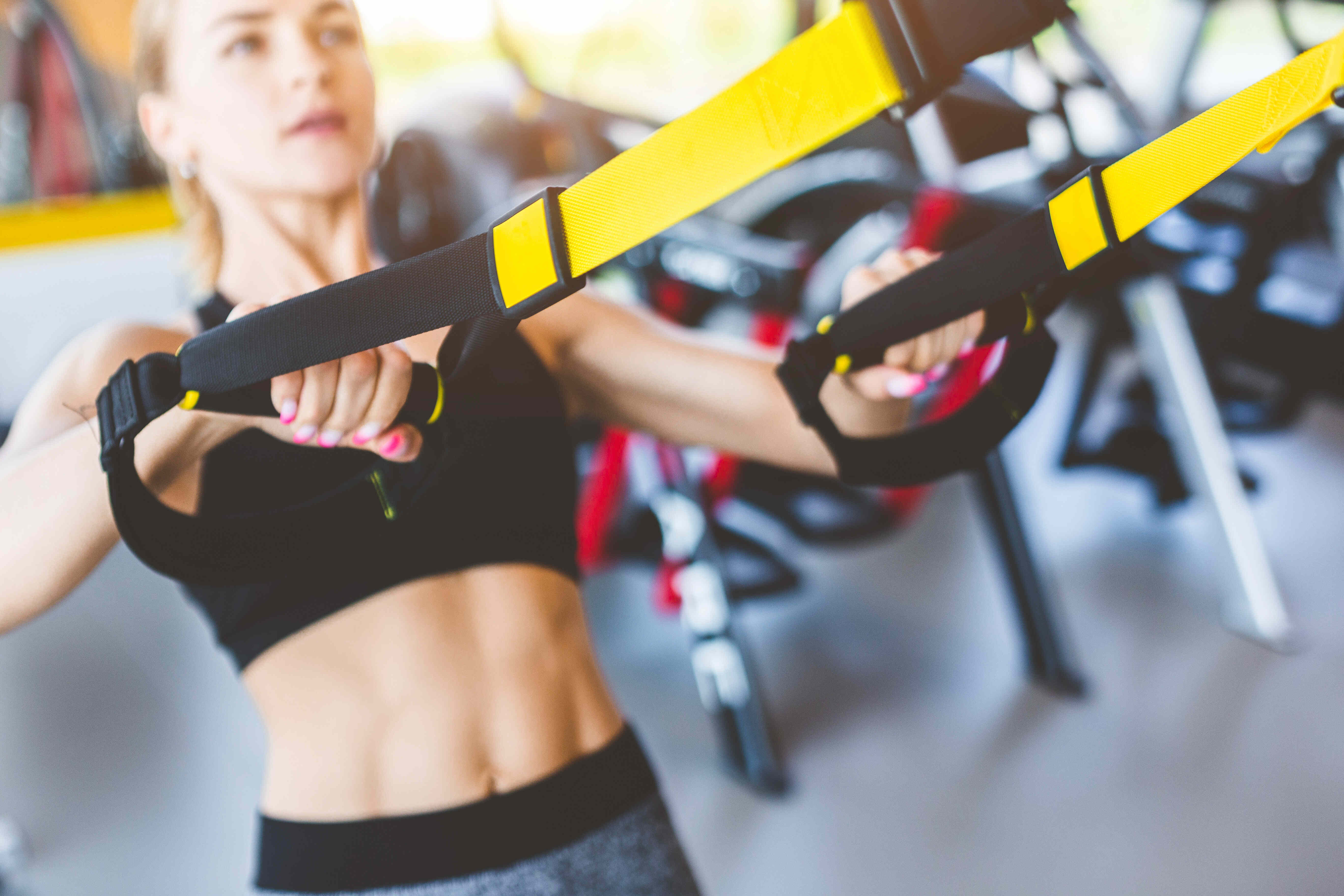 Rabenberg Sportkurse: Sling Training Das Bild zeigt eine Frau beim Training im Fitnessstudio. Sie hält sich mit beiden Händen an gelben und schwarzen Schlaufen fest. Ihr Blick ist nach oben gerichtet, und sie wirkt konzentriert. Die Frau trägt Sportkleidung. Im Hintergrund sind weitere Trainingsgeräte zu sehen, wobei die Umgebung unscharf dargestellt ist.