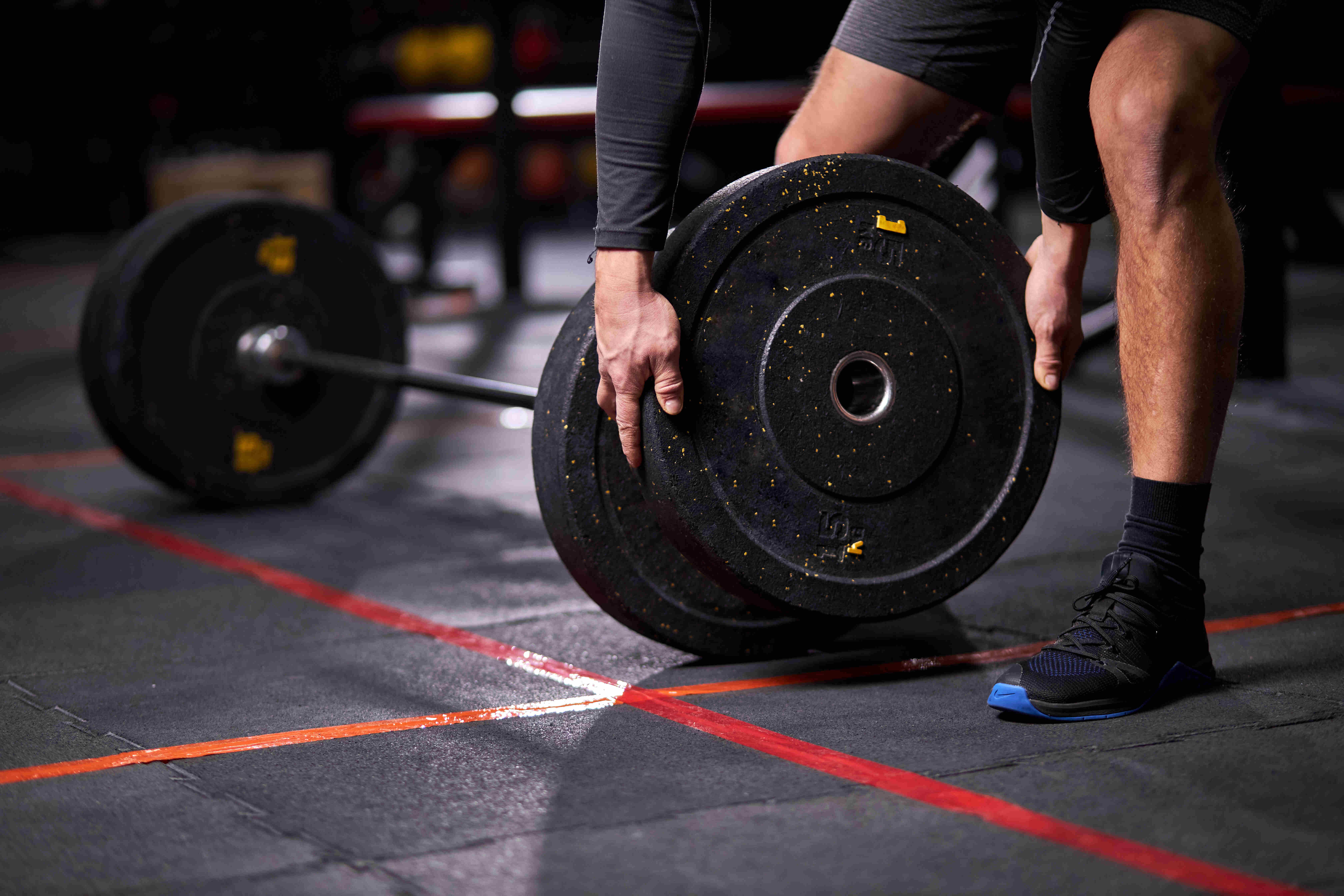 Rabenberg Sportkurse: Heavy-Cross Das Bild zeigt eine Person, die Gewichte in einem Fitnessstudio anhebt. Die Person hält eine schwarze Gewichtsscheibe mit beiden Händen. Der Körper der Person ist angeschnitten, aber man kann sehen, dass sie schwarze Kleidung trägt. Auf dem Boden sind rote Linien zu sehen. Der Hintergrund ist unscharf, was darauf hindeutet, dass der Fokus auf der Person und den Gewichten liegt.