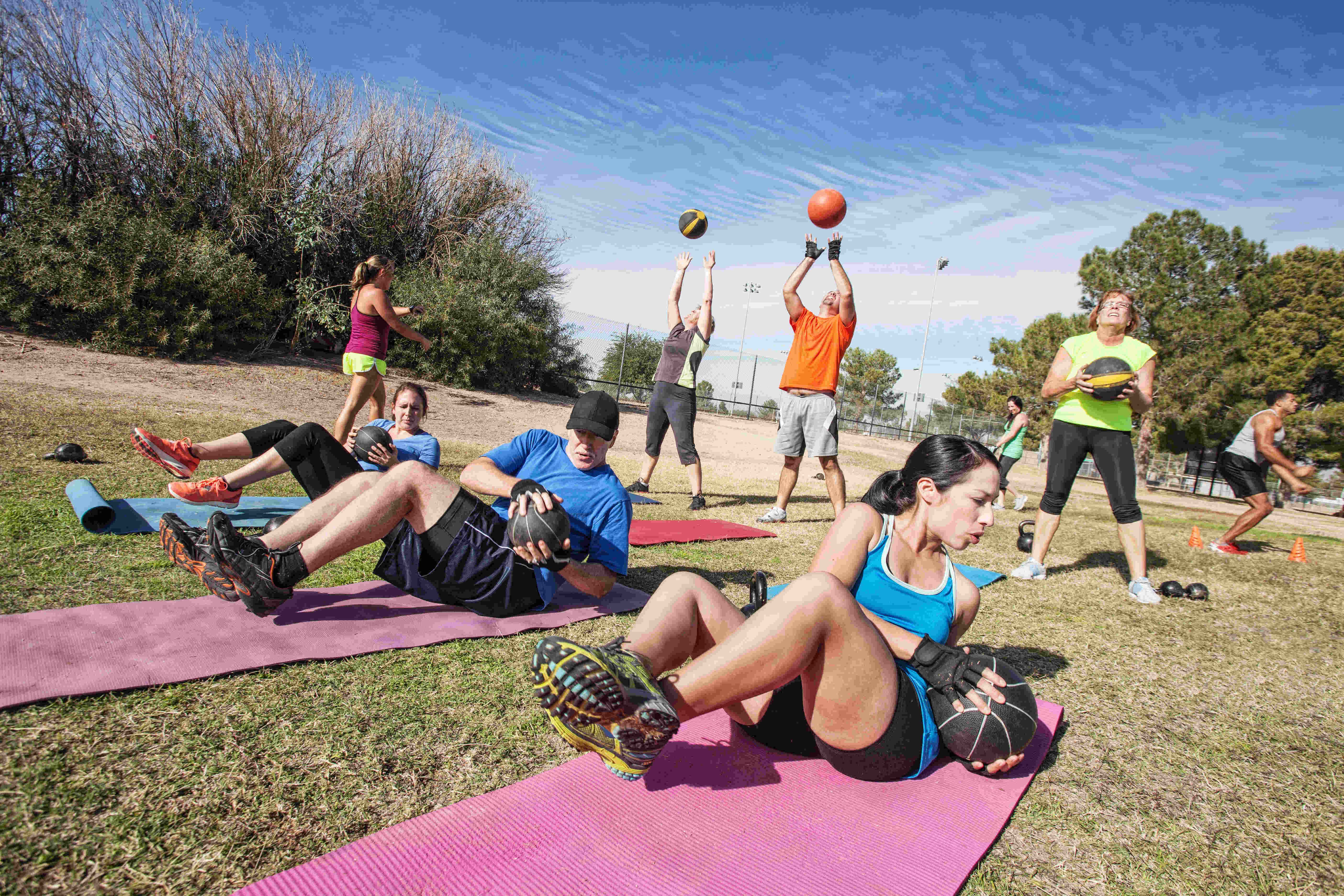 Rabenberg Sportkurse: Outdoor-Cross Das Bild zeigt eine Gruppe von Menschen, die im Freien trainieren. Einige liegen auf Matten und machen Bauchmuskeltraining mit Medizinbällen. Andere heben die Bälle in die Luft oder halten sie. Im Hintergrund sind Bäume und blauer Himmel zu sehen.
